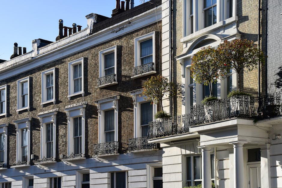A row of classic London townhouses with brick facades and white window frames, each featuring small decorative wrought-iron balconies. The balconies are adorned with potted plants and small trees with round, leafy canopies, adding greenery to the urban setting. The buildings are illuminated by sunlight, casting shadows on the facades, and the sky above is clear and blue, emphasizing a bright, crisp day. The scene highlights polished architectural details and well-maintained plantings, reflecting the outdoor maintenance and landscaping work typical for Westminster office estates and residential blocks, as handled by Gardeners Westminster.