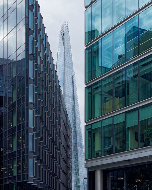 A view looking up between modern office buildings with glass facades, reflecting the surrounding structures and the cloudy sky above. The building on the left has a dark, textured exterior with rectangular glass panels arranged in a grid pattern, while the building on the right features large, horizontal glass windows with visible interior furnishings and potted plants on some floors. In the background, the slender, pointed Shard skyscraper dominates the skyline, its glass surface catching the light. The scene depicts an urban landscape emphasizing sleek architecture, clean lines, and the reflective qualities of contemporary glasswork typical of Westminster's commercial district, with no visible gardening or outdoor maintenance activities. The focus is on the architectural environment rather than landscaping, highlighting the city's modern skyline.