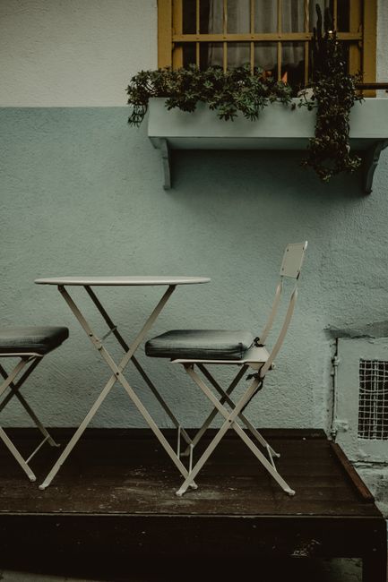 A small urban garden terrace with a weathered wooden deck floor, featuring a round, white metal bistro table and two matching folding chairs, one with a black cushioned seat and the other partially visible. Behind the seating area, a pale grey wall with a decorative window has a window box filled with lush, dark green foliage and trailing plants. The scene is illuminated by natural daylight, highlighting the textures of the chipped paint on the wall, the rough wooden decking, and the vibrant greenery. This outdoor space, managed by Gardeners Westminster, exemplifies compact garden solutions suitable for terraces and courts, emphasizing garden maintenance and landscaping work that enhances small garden areas in urban settings.